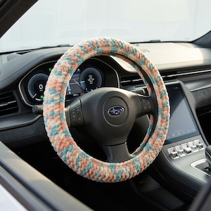 May include: A thick, crocheted steering wheel cover featuring a variegated yarn pattern in soft shades of peach, light blue, and cream. The textured, plush accessory is fitted snugly around the circular rim of a black Subaru steering wheel.