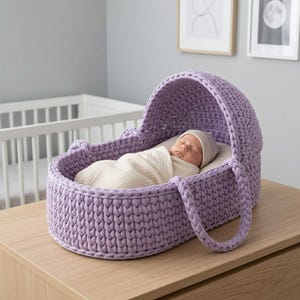 Könnte beinhalten: Ein lavendelfarbener Babykorb aus grobem Strick mit passender Haube und zwei seitlichen Tragegriffen. Im Inneren liegt ein Baby mit beiger Mütze, eingewickelt in eine cremefarbene Decke, auf einer glatten, cremeweißen Matratzenauflage.