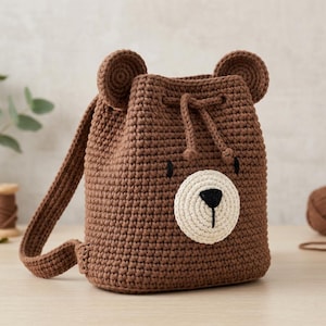 May include: A brown crochet backpack featuring a bear face design with two rounded ears on top, a cream-colored circular snout, a black embroidered nose and mouth, and a drawstring closure with two matching brown cord ties and shoulder straps.