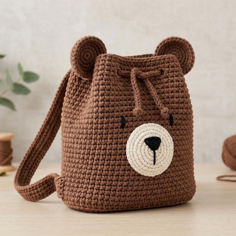 May include: A brown crochet backpack featuring a bear face design with two rounded ears on top, a cream-coloured circular snout, a black embroidered nose and mouth, and a drawstring closure with two matching brown cord ties and shoulder straps.