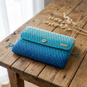 Könnte beinhalten: Eine rechteckige Häkeltasche mit einem Farbverlauf von Hellblau an der oberen Klappe bis zu einem tiefen Marineblau an der Basis. Die Klappe ist mit einem runden goldfarbenen Knopf und einem kleinen rechteckigen Metall-Markenschild versehen.