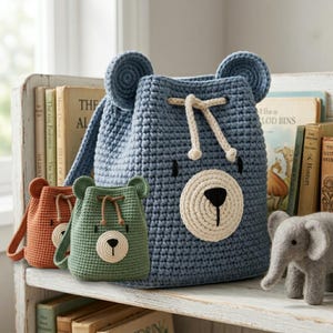 May include: A large blue crochet backpack with bear ears and a cream-coloured muzzle, accompanied by a smaller green crochet bear bag and a rust-coloured crochet bear bag. A small grey felt elephant figurine stands to the right of the bags.