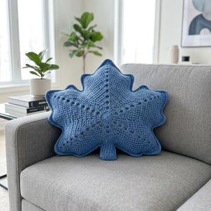 May include: A blue crochet pillow shaped like a maple leaf. The pillow features a textured, hand-stitched surface with a central vein pattern created by open-work stitches radiating from the center toward the pointed edges of the leaf shape.