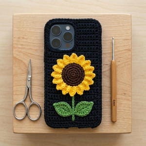 May include: A dark navy blue crochet phone case featuring a three-dimensional sunflower motif with bright yellow petals, a dark brown centre, and two green leaves. The case is positioned next to a pair of silver scissors and a 4mm wooden crochet hook.