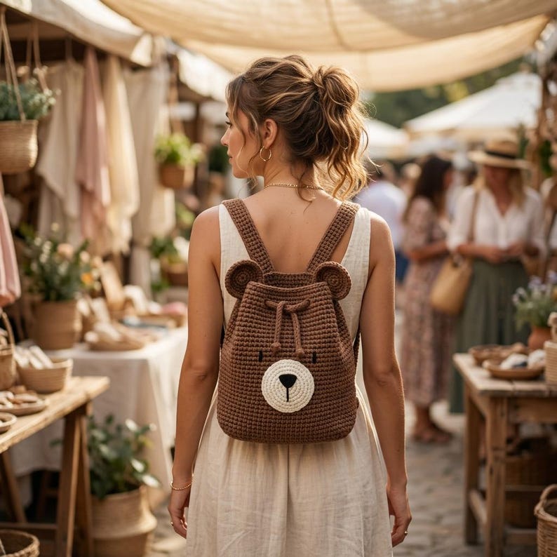 May include: A brown crochet backpack featuring a bear face design with small rounded ears, a cream-coloured muzzle, and a black nose. The bag has a drawstring closure and two shoulder straps, worn over a simple sleeveless cream-coloured dress.