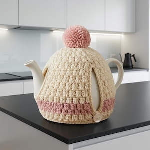 May include: A white ceramic teapot covered by a cream-colored crocheted cozy featuring a textured bobble stitch pattern, a dusty pink pom-pom on top, a horizontal dusty pink stripe near the base, and a single white button closure at the side.