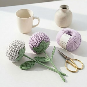 Könnte beinhalten: Zwei gehäkelte Blumen mit strukturierten hellgrauen und lavendelfarbenen Blütenblättern, grünen Stielen und Blättern. Daneben stehen eine cremefarbene Keramiktasse, eine kleine beige Vase, ein Knäuel lavendelfarbenes Baumwollgarn mit Etikett und eine goldfarbene Schere.