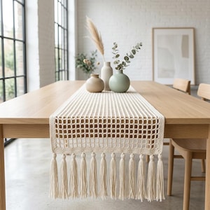 May include: A cream-colored, open-weave table runner with a grid pattern and long tassels. The runner is displayed on a light wood dining table, complemented by decorative vases and framed artwork. The room has large windows and a neutral color palette.
