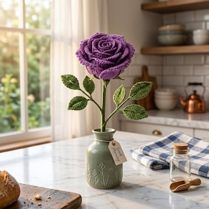May include: A crocheted purple rose with green leaves and a stem, displayed in a light green ceramic vase. A small rectangular paper tag tied to the vase neck features the printed text Good Morning in black ink, adding a simple detail.