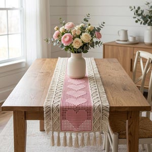 May include: A rectangular table runner featuring a central pink crocheted panel with a repeating heart pattern, bordered by cream-colored macrame detailing and finished with long fringe tassels on both ends, displayed on a wooden table.