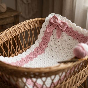 May include: A handmade crochet blanket featuring a white and soft pink shell stitch pattern. The blanket is rectangular with a scalloped white border and a prominent, matching pink crocheted bow attached to the top corner of the fabric.