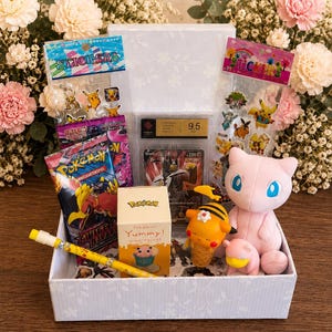 Pode incluir: Uma caixa de presente branca contendo uma carta Pokemon graduada, duas folhas de adesivos, um pacote de cartas Pokemon, uma caneta amarela estampada, uma pequena caixa Pokemon Yummy, um boneco Pikachu abelha e uma pelúcia rosa do Mew.