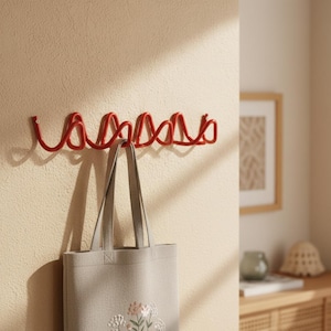 May include: A red metal wall hook with a looping design is mounted on a beige wall. A gray tote bag with floral embroidery hangs from one of the hooks. A framed picture and a wooden cabinet are in the background.