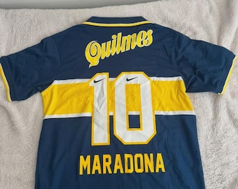May include: A navy blue soccer jersey featuring a horizontal yellow stripe across the chest. The back displays the text Quilmes in yellow, the number 10 in white with yellow trim, and the names Maradona and Xeneizes in yellow lettering.