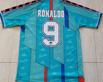 May include: A light blue short-sleeve soccer jersey featuring the name RONALDO and the number 9 in white on the back. The shirt has a repeating pattern of the word Barca and the Kappa logo, with black and red striped trim along the sleeves.