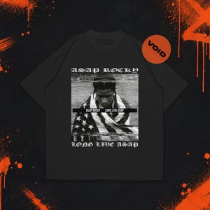 Puede incluir: Una camiseta negra con un estampado gráfico blanco de A$AP Rocky envuelto en una bandera estadounidense. El texto "A$AP ROCKY" aparece sobre la imagen, "LONG LIVE ASAP" debajo y "A$AP ROCKY LONG.LIVE.A$AP" en el centro. Se ve un pequeño círculo naranja con "VOID".