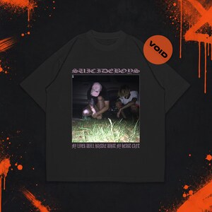 Pode incluir: Uma t-shirt preta de manga curta com uma impressão gráfica de duas pessoas agachadas numa zona relvada à noite. O texto "SUICIDEBOYS" aparece acima da imagem e o texto "MY LIVER WILL HANDLE WHAT MY HEART CANT" está impresso abaixo.