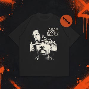 Puede incluir: Una camiseta negra de manga corta con un estampado gráfico blanco de alto contraste de A$AP Rocky. El diseño muestra un retrato con manos tirando de los párpados, un gráfico de un coche pequeño y el texto A$AP ROCKY. Se observa un logotipo circular naranja VOID.