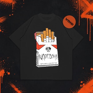 Pode incluir: Uma t-shirt preta com um gráfico de um maço de cigarros branco com detalhes em laranja. O maço exibe um ícone de caveira e o texto "SUICIDEBOY$" numa fonte estilizada. Um pequeno círculo laranja com a palavra "VOID" está posicionado no canto superior direito.
