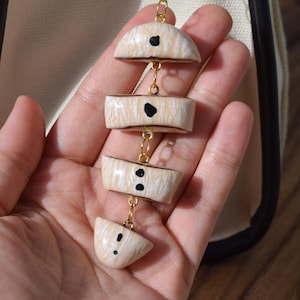 May include: A vertical pendant ornament featuring four cream-coloured, semi-circular segments with a wood-grain texture and black dot accents. The segments are connected by small gold-toned metal jump rings in a linear arrangement.