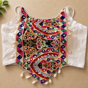Peut inclure: Un haut blanc sans manches doté d'un panneau central avec une broderie multicolore vive, de petits miroirs circulaires et des coquillages cauris blancs suspendus le long des bords et de l'ourlet pointu, créant un aspect détaillé et texturé.