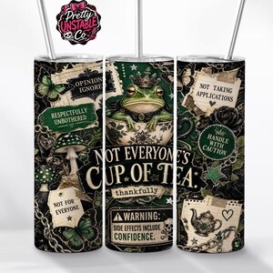 May include: A cylindrical tumbler with a metal straw featuring a dark green and black collage design. It displays a frog wearing a crown in a teacup, mushrooms, butterflies, chains, and text including "Not Everyone's Cup of Tea" and "Warning: Side Effects Include Confidence."