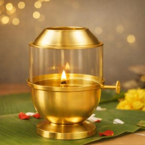 May include: A gold-toned metal oil lamp featuring a cylindrical glass chimney, a rounded base, and a small side adjustment knob. The lamp has a lit wick centered within the glass enclosure, casting a warm glow from the flame inside the metal structure.