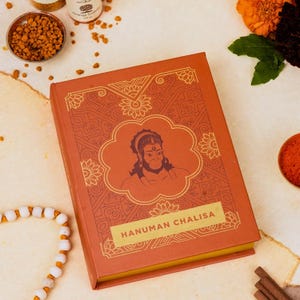 May include: A rectangular book titled Hanuman Chalisa with a burnt orange cover featuring gold-colored line art of a deity in a central cloud-shaped frame, surrounded by intricate geometric and floral patterns, with a yellow spine edge visible.