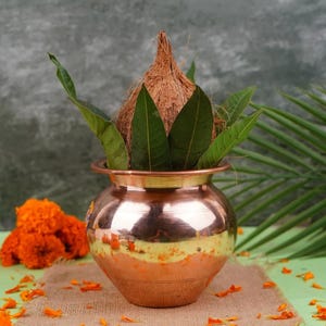 May include: A polished copper pot with a rounded body and a wide rim, containing a brown fibrous coconut nestled among several green mango leaves. The pot sits on a textured brown mat surrounded by scattered bright orange marigold flower petals.