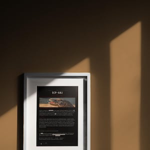 May include: A white framed print featuring a black document with the text SCP-682 at the top, followed by a photograph of a reptile head and several paragraphs of text with redacted sections, presented in a clean, rectangular wall frame.