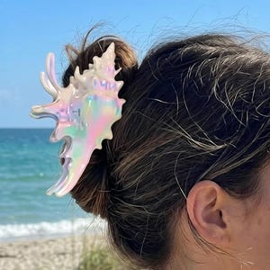 Könnte beinhalten: Eine Haarklammer in Form einer Muschel mit irisierendem, perlmuttartigem Finish, das Licht in Rosa-, Blau- und Weißtönen reflektiert. Die Klammer ist in braunem Haar befestigt und zeigt ein detailliertes, stacheliges und organisches Spiralmuscheldesign.