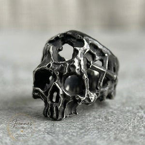 May include: A dark silver-toned ring featuring a highly textured, weathered skull design with hollowed-out eye sockets and nasal cavity. The band incorporates cross-shaped cutouts on the sides, finished with a distressed, oxidized metal patina.