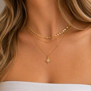 Op de afbeelding: Een goudkleurige gelaagde ketting bestaande uit drie afzonderlijke kettingen. De bovenste ketting heeft platte ronde schakels, de middelste ketting heeft kleine kraaltjes en de onderste ketting heeft een stervormige hanger met een witte steen.