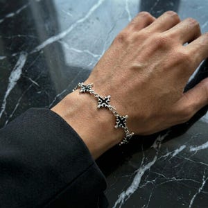 May include: A silver chain bracelet featuring repeating cross-shaped links with black enamel detailing. The bracelet is constructed from polished metal links that connect each decorative cross element, creating a continuous pattern around the wrist.