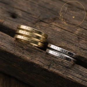 May include: Three gold-toned rings stacked together, each engraved with the names EMMA, GRAYSON, and CHLOE, alongside two silver-toned rings stacked together, each engraved with the names FELICITY and HARRIS, all featuring a smooth, flat band design.