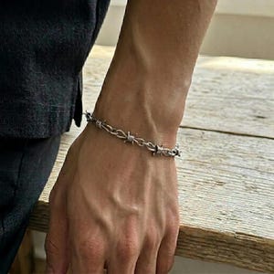 Könnte beinhalten: Ein Armband aus silberfarbenem Metall mit einem Design, das Stacheldraht nachempfunden ist. Die Kette besteht aus gedrehten Metallgliedern mit kleinen, spitzen Vorsprüngen, die gleichmäßig über die gesamte Länge verteilt sind.