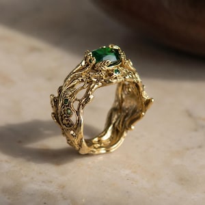 Op de afbeelding: Een gouden ring met een complexe, getextureerde band die doet denken aan verstrengelde takken en bladeren. De ring is in het midden gezet met een opvallende, gefacetteerde smaragdgroene edelsteen, geaccentueerd door diverse kleine, ronde groene stenen.