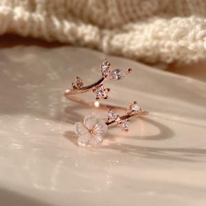 Puede incluir: Un anillo abierto de oro rosa con diseño de rama, adornado con piedras de circonita cúbica transparentes y una flor blanca. El anillo es ajustable y presenta un delicado trabajo en metal con piedras brillantes a lo largo de los tallos.