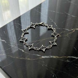 Puede incluir: Pulsera plateada con un diseño de cruces repetidas. La pulsera está compuesta por eslabones en forma de cruz interconectados, creando un patrón delicado e intrincado. El cierre es visible, y la pulsera descansa sobre una superficie oscura, similar al mármol.