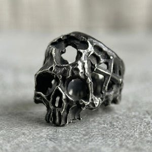 May include: A dark silver-toned metal ring featuring a highly textured, weathered skull design with hollowed-out eye sockets and nasal cavity, accented by a cross symbol integrated into the side of the band with a rugged, oxidized finish.