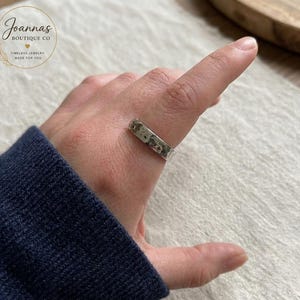 Puede incluir: Un anillo de banda color plata con un acabado martillado y texturizado que se lleva en un dedo. El anillo presenta un perfil plano y un brillo metálico, mostrando un patrón de superficie irregular y abollado en toda la circunferencia.