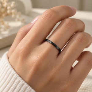 Può includere: Un anello sottile di colore nero con una finitura superficiale martellata e testurizzata. La fascia presenta un aspetto scuro e opaco con sottili rientranze sull'esterno, creando un motivo distinto e irregolare lungo tutta la circonferenza.
