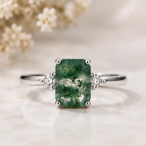 May include: A silver ring featuring a rectangular emerald-cut moss agate gemstone with green inclusions. The center stone is flanked by two small, round, clear accent stones set on a polished silver band with four prongs holding the main gem.