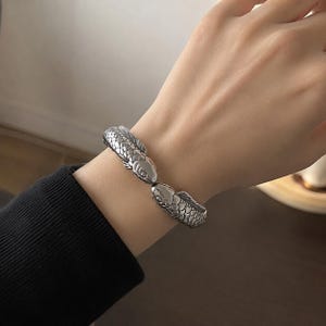 以下が含まれることがあります： 中央で2匹の鯉の頭が向き合うデザインのシルバーカラーのメタル製カフブレスレット。ブレスレット全体には細かく質感のある鱗模様が施されており、魚の頭が向かい合って留め具のような形状を形成しています。