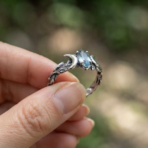 May include: A silver ring featuring a marquise-cut light blue gemstone set between two crescent moon shapes. The band is crafted with a detailed, textured vine and leaf pattern that wraps around the finger, creating a nature-inspired design.