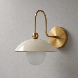 May include: A wall-mounted light fixture featuring a circular brass wall plate, a curved brass arm, and a glossy cream-colored dome shade. A spherical frosted white glass bulb is positioned beneath the shade, centered on a small brass ball connector.