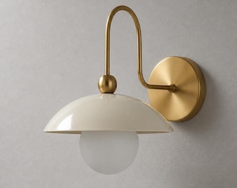 May include: A wall-mounted light fixture featuring a circular brass wall plate, a curved brass arm, and a glossy cream-colored dome shade. A spherical frosted white glass bulb is positioned beneath the shade, centered on a small brass ball connector.