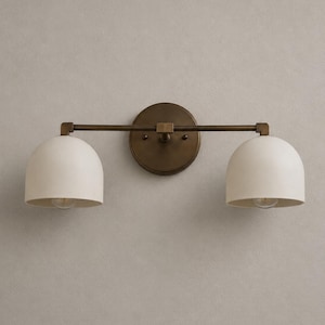 May include: A two-light wall sconce featuring a circular antique brass backplate and a horizontal bar supporting two matte off-white dome-shaped shades. The fixture has a metallic finish on the central mounting plate and the connecting hardware.