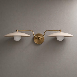 May include: A double wall sconce featuring a central circular brass mounting plate with two extending arms. Each arm supports a wide, shallow, off-white saucer-shaped shade positioned above a small, spherical, frosted white glass light bulb.