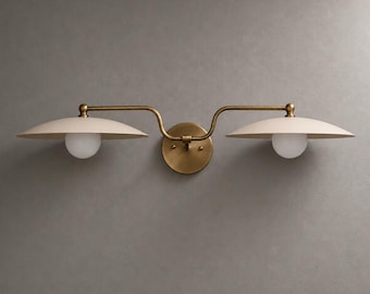 May include: A double wall sconce featuring a central circular brass mounting plate with two extending arms. Each arm supports a wide, shallow, off-white saucer-shaped shade positioned above a small, spherical, frosted white glass light bulb.